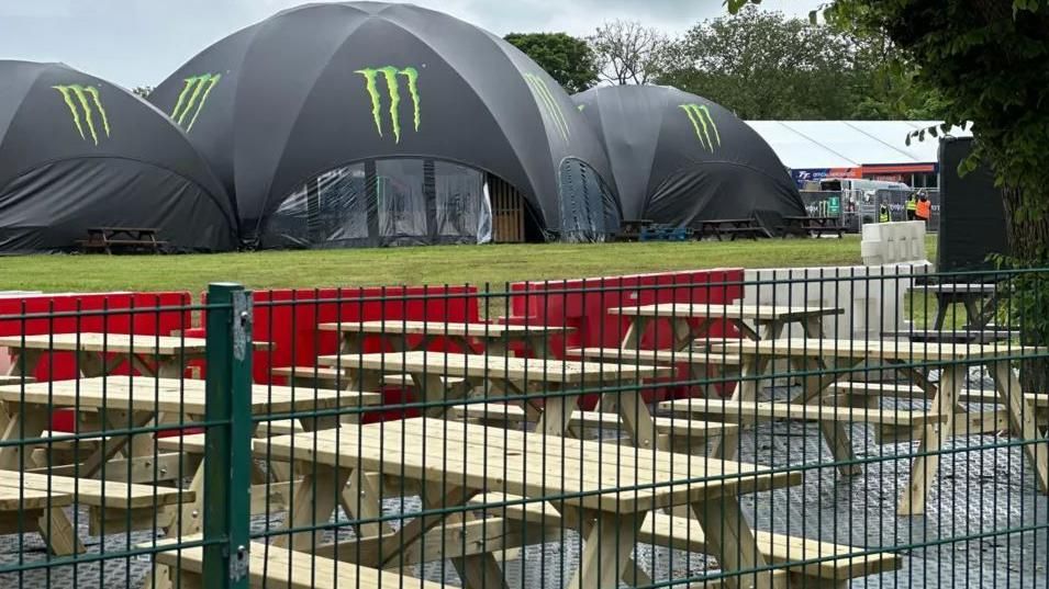 A series of wooden picnic tables and benches laid out in front of three big black tents with the Monster Energy logo on the front. Tables and tents are behind a green fence in the fan zone area in Noble's Park.