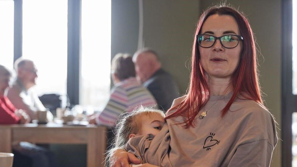 Lottie Middlemass breastfeeding her daughter Quinn underneath her jumper at a cafe with people in the background, wearing black glasses with red hair