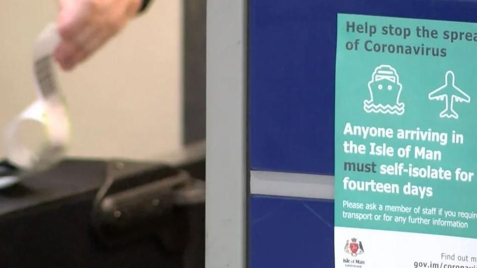 A sign says anyone arriving in the Isle of Man must self-isolate for fourteen days, to stop the spread of coronavirus. The sign is green and white. In the background there's a blurred out suitcase and a hand holding a lable.