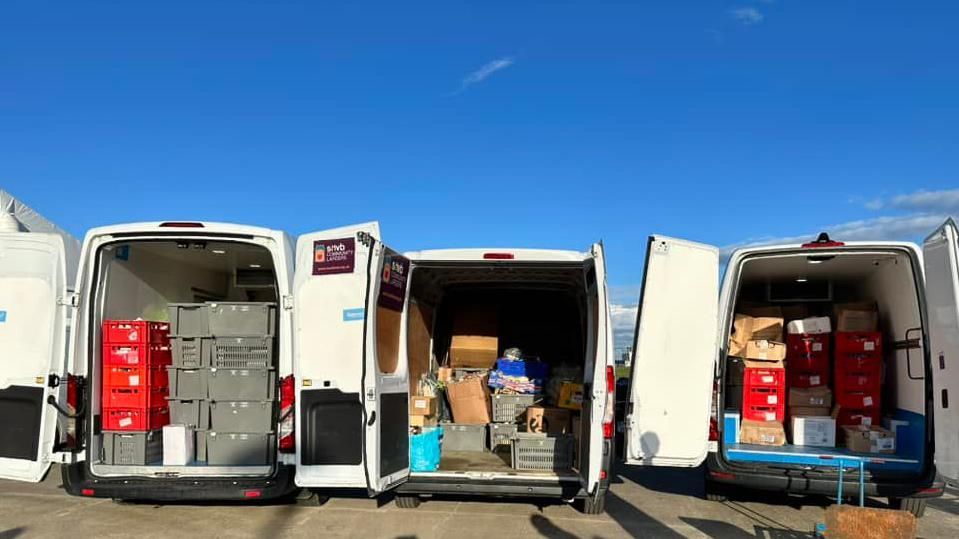 Three white vans with back doors open revealing crates of food