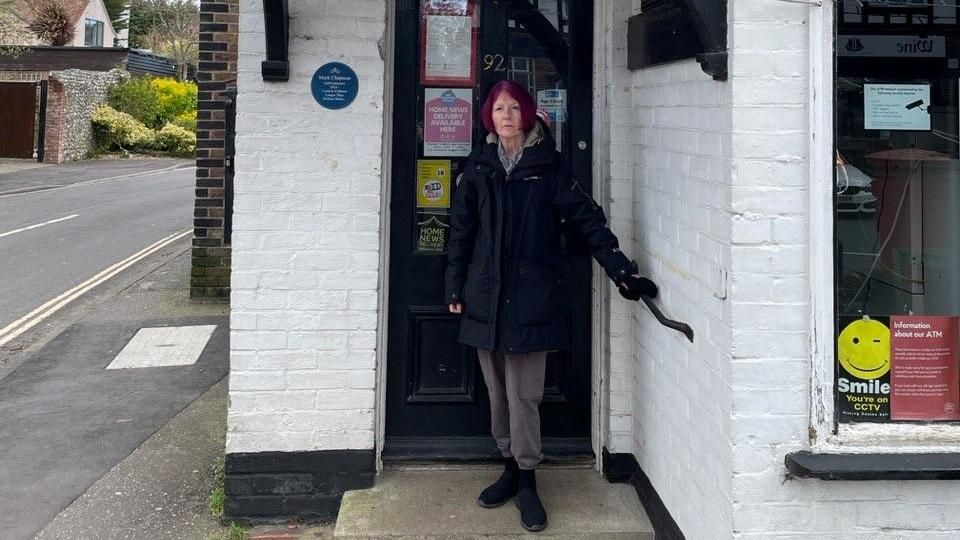 A woman with short purple hair poses outside the white brick post office building in Felpham in a dark hooded parka style coat