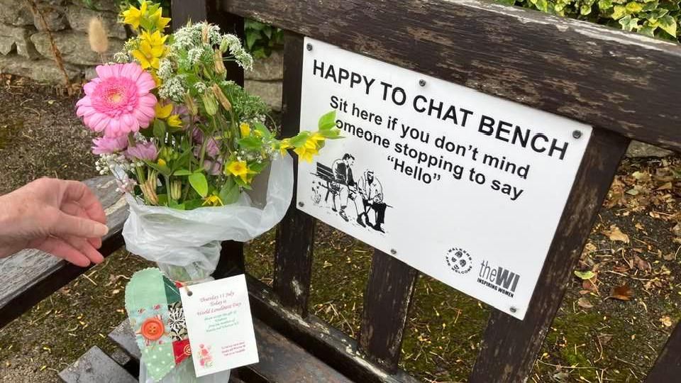 Somebody placing a bunch of flowers on the corner of a wooden bench. The flowers are tied with a green patchwork cloth heart and note. On the bench is a sign which says it is a: "Happy to chat bench".