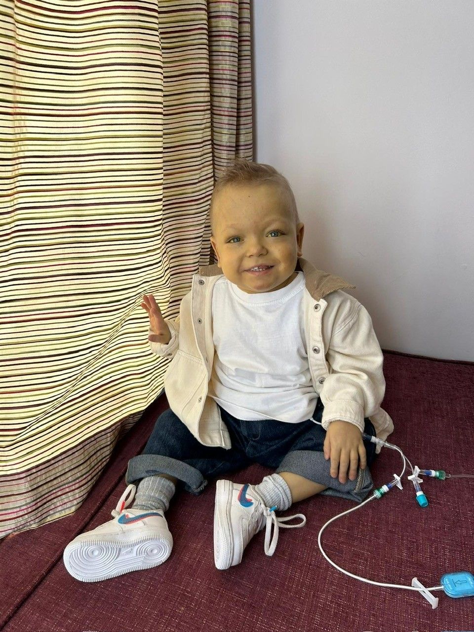 Zachary sits on the carpet beside a stripy curtain, his medical tubes visible beside him