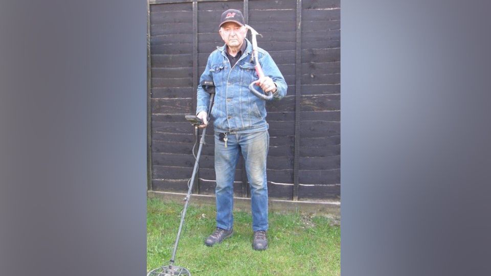 Tom Clarke in double denim and holding his metal detector in his right hand, his spade in his left