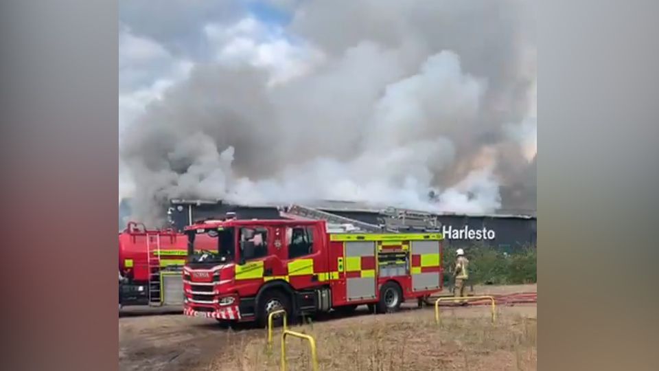 A fire engine is parked in front of a warehouse with large plumes of smoke rising from the roof