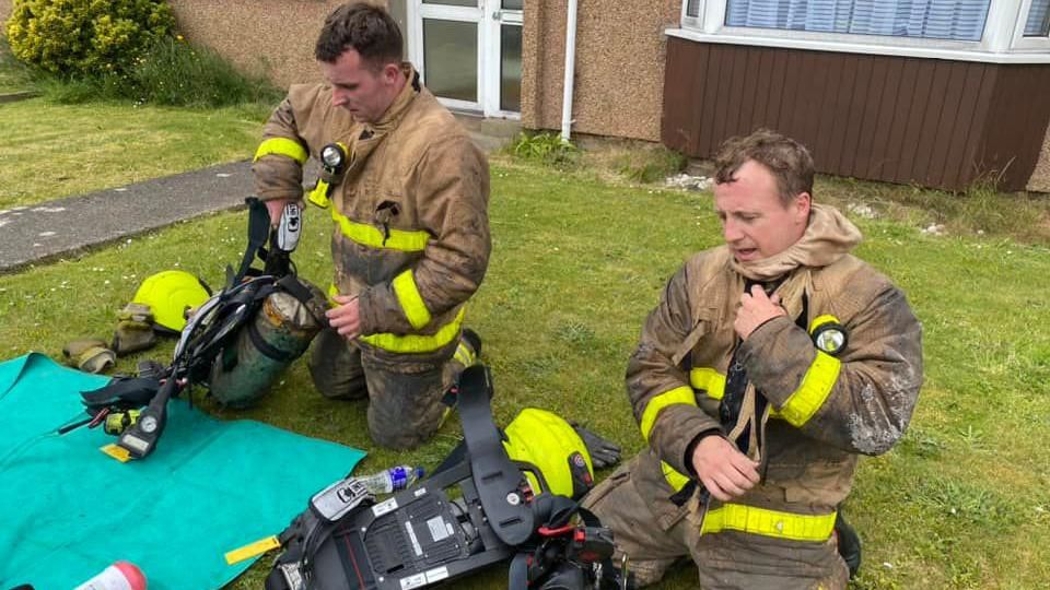 Two firemen remove their equipment in a garden. 