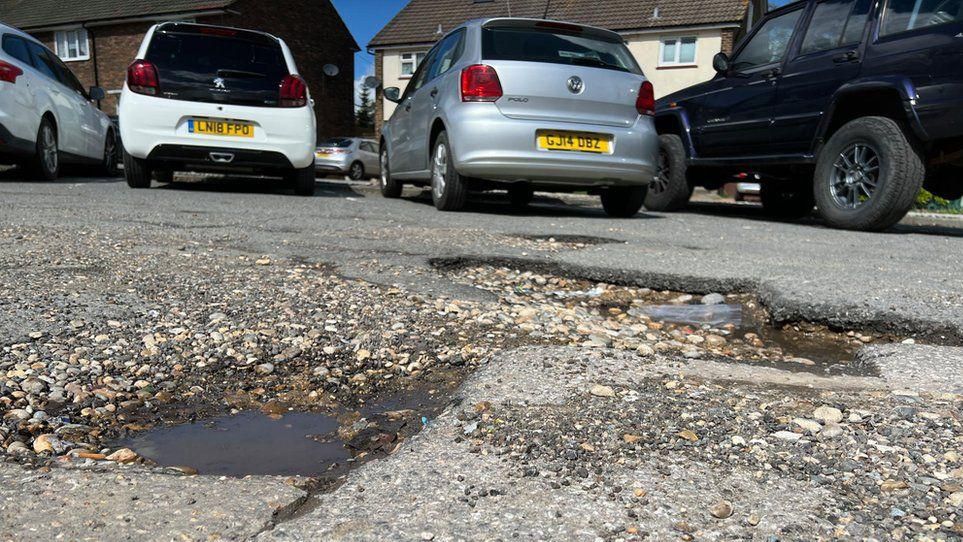 A potholed road with parked cars in the background