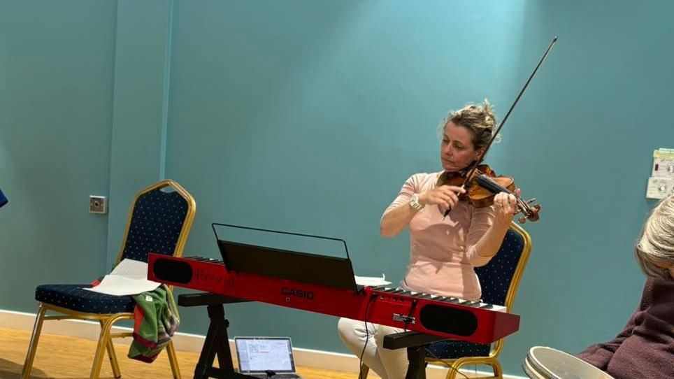 Caroline Speca plays the violin as she is sitting in front of a red keyboard. She is wearing a pink top and white trousers and is looking down at a sheet of music. 