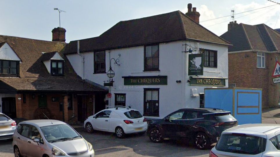 Street view of the pub with a number of cars parked next to it. It is a white building with a green sign with 'Chequers' written on it in gold embossed lettering.