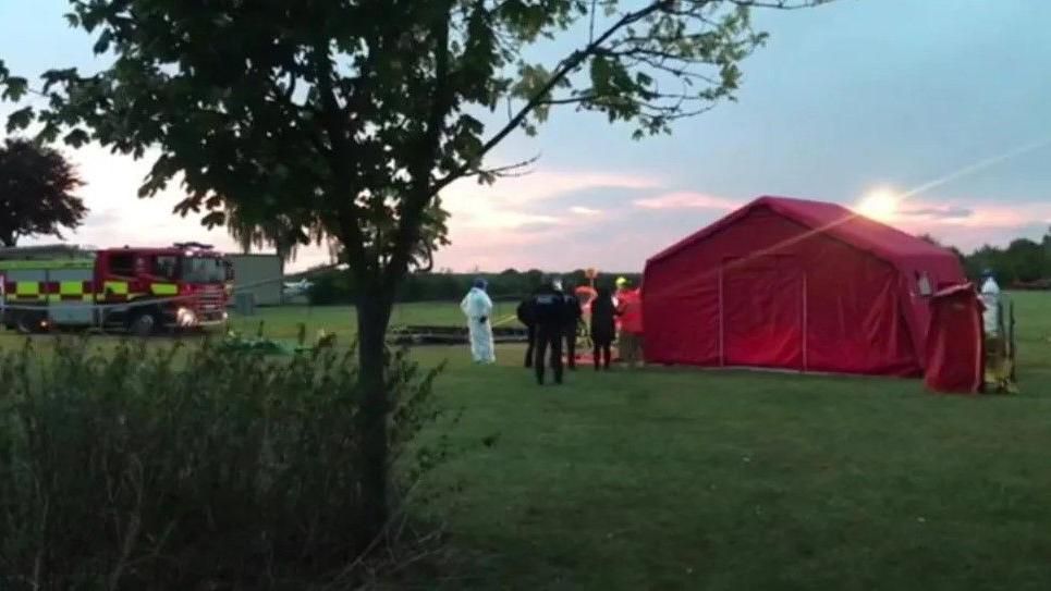 A fire engine and a large red tent can be seen in a field with emergency service workers stood alongside it. There is a small tree and some shrubbery in the foreground.