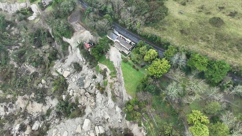 Large rocks and boulders strewn down cliff side buildings and road among trees 