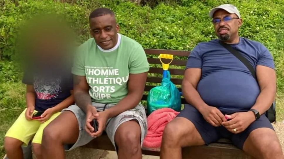 Siyabonga Twala sat on a bench with his family. He wears a green t-shirt and grey shorts.