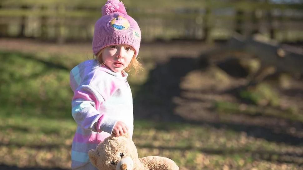 Aoife, a three year old girl, wears a pink bobble hat and clutches a teddy bear by the ear.