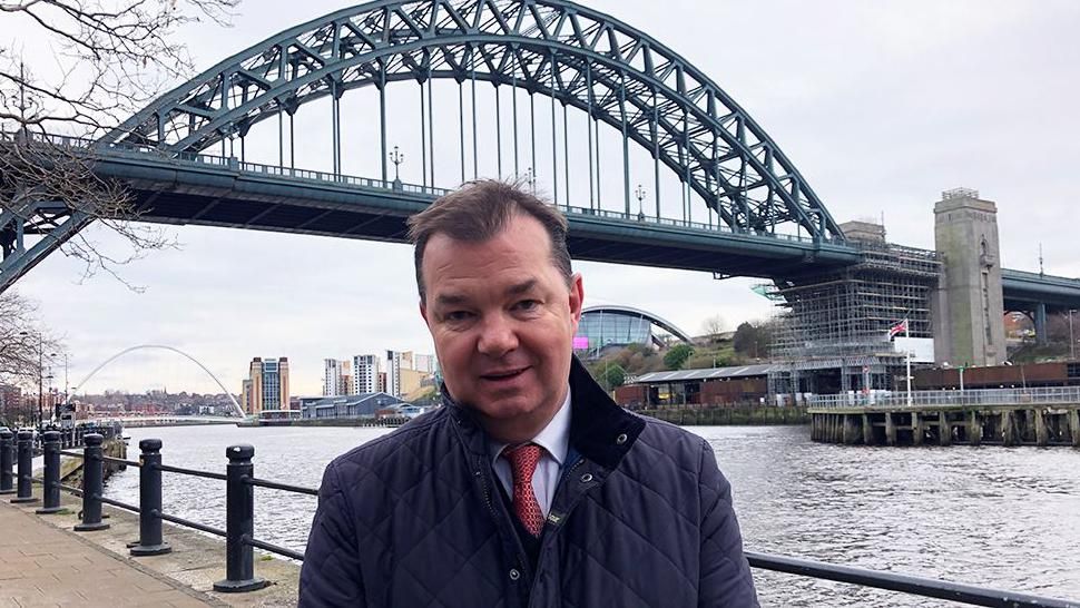 Guy Opperman standing on Newcastle Quayside with Gateshead and the Tyne Bridge behind him