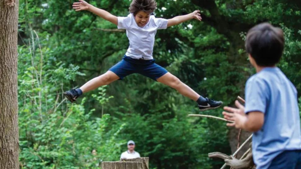 A boy doing a star jump off some play equipment in the arboretum