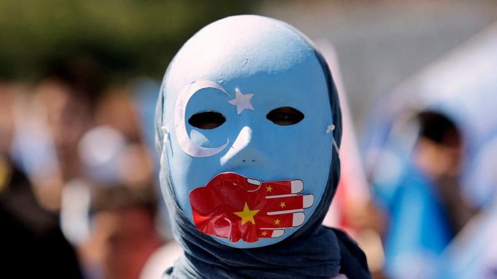 A Uyghur protester wearing a blue mask decorated with a star and crescent moon symbol, with a red hand bearing the Chinese flag covering the mouth