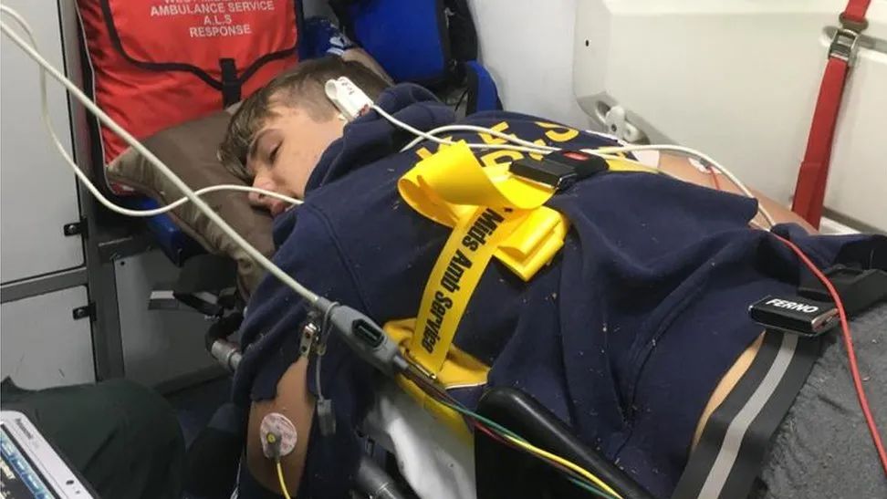 A man lies face down on an ambulance stretcher, with a yellow securing belt around his back