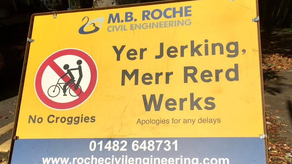 A yellow road sign featuring a joke traffic symbol showing a bicycle rider giving a passenger a "croggy" and black letters reading "Yer Jerking, Merr Rerd Werks" (you're joking, more road works)