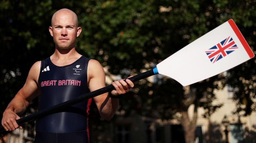 Sam Murray in his Great Britain rowing gear holding a white ore with a Union Jack on it