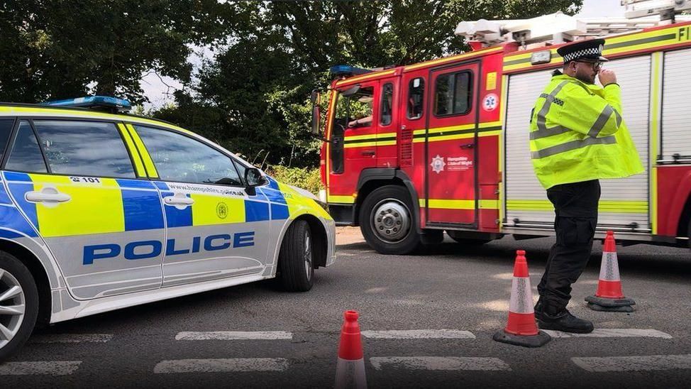 Fire engine right and police car left with noses of both vehicles meeting in the centre - police officer standing between cones