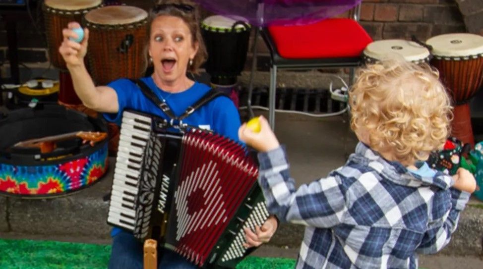 A woman working for the charity, with blonde hair and a blue t-shirt, plays a red accordion with a child who has blonde, curly hair