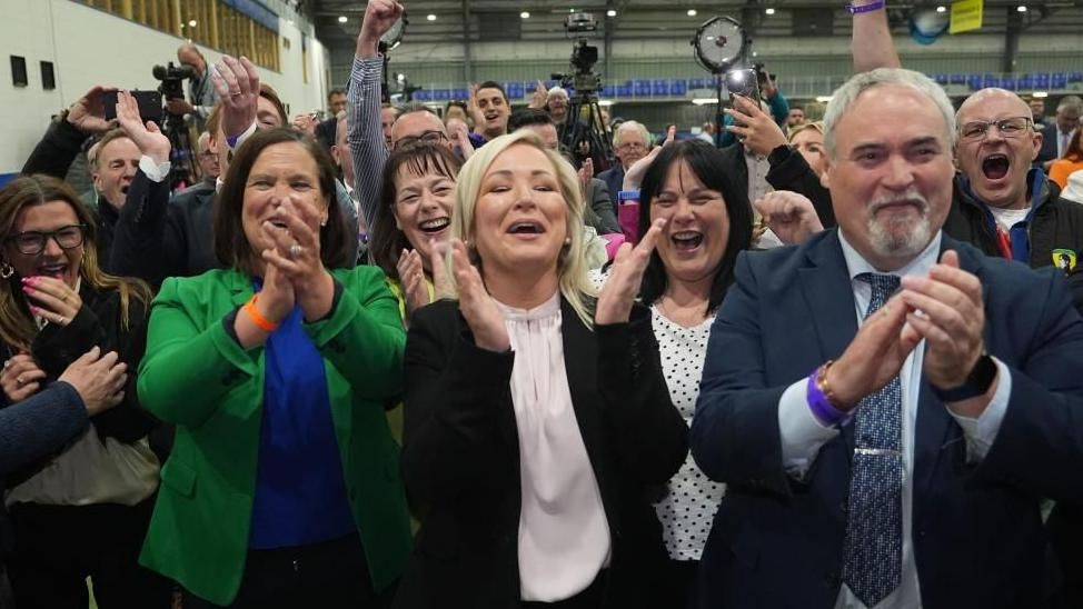 Michelle O'Neill  clapping alongside a crowd of jubilant party members 