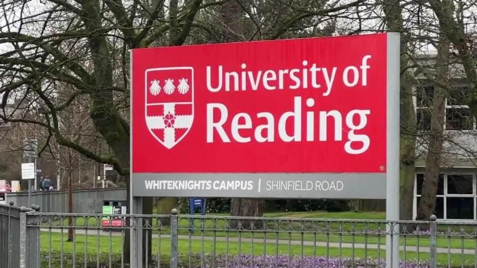 Red University of Reading sign outside campus building. There are purple flowers under the sign in front of an area of grass. There are four trees behind with no leaves, they are in front of a white building with large windows.