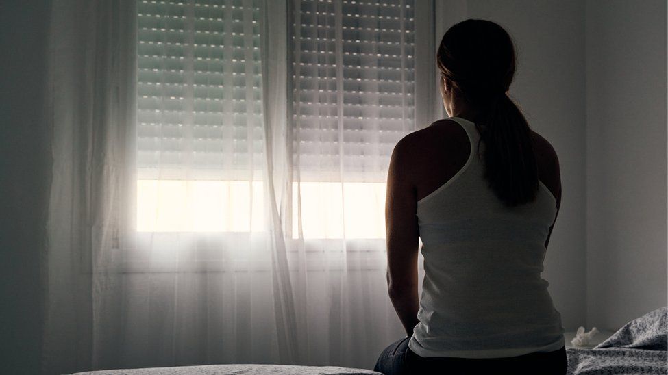 A woman sits on a bed with her back to the camera looking at a window.