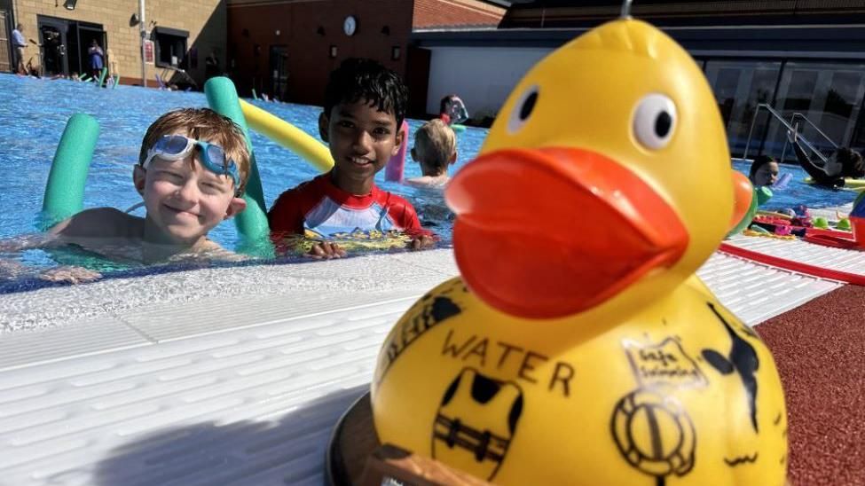 Two boys on woggles in the outdoor pool smiling at the side behind a large yellow rubber duck with photos of lifejackets and boys and divers on it