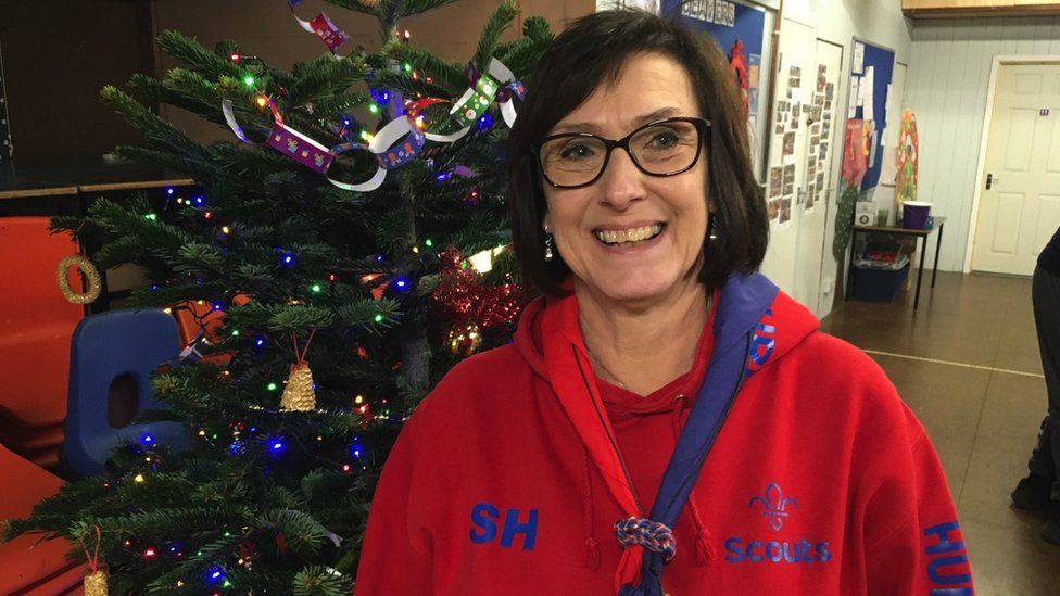 Sue Hunter. She is wearing a red hoodie with SH on one side. She is stood in front of a Christmas tree and is looking directly at the camera and smiling.