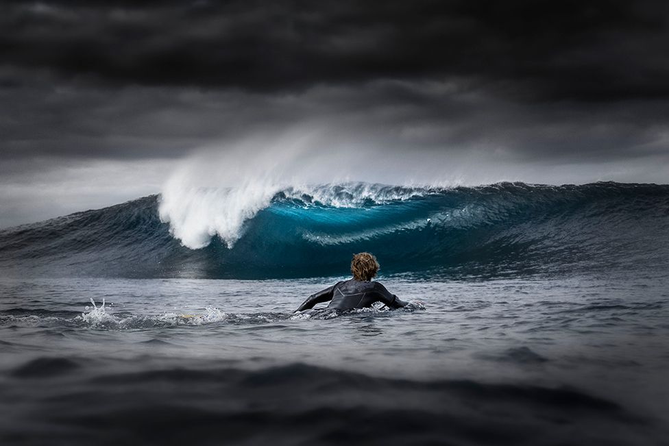 A bodyboarder paddles out to sea, while big storm clouds block the sun and create an electric blue colour effect on the wave.