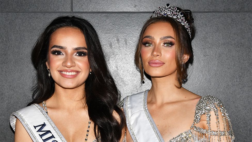 UmaSofia Srivastava and Noelia Voigt looking at the camera, they are both wearing sparkly dresses, sashes and Noelia is wearing a crown