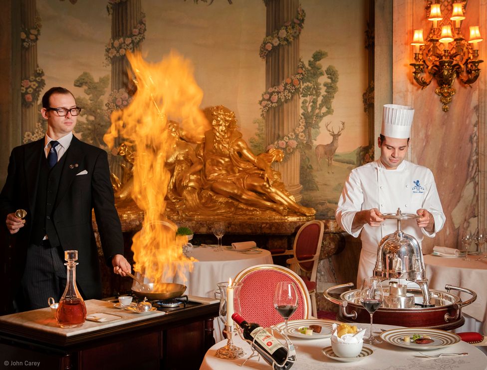 Flames leap from a pan used by a man who is stood at a small cooker next to a set table, while a second man in chef whites uses a metal contraption