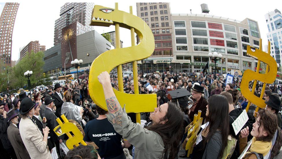 Молодой человек держит знак доллара во время митинга «Захвати Уолл-стрит» против высокой стоимости обучения в колледже 25 апреля 2012 года в Нью-Йорке. Множество студентов и бывших студентов собрались, чтобы пожаловаться на высокую стоимость обучения и ссуд для колледжа. 25 апреля ознаменовался днем, когда студенческий долг США достиг 1 триллиона долларов