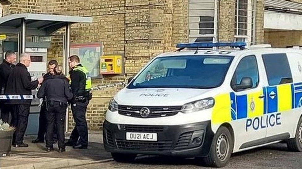 Two police cars parked outside the Harwich Town railway station. There is a police cordon and a group of people standing behind the police cars. One male officer is standing in front.