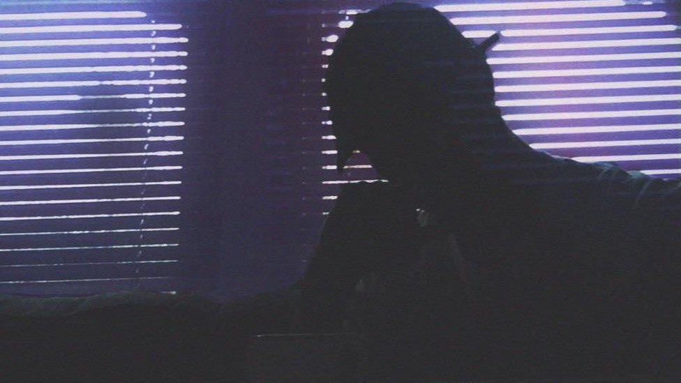 A gloomy photo of a man silhouetted against two windows with slatted blinds across them.