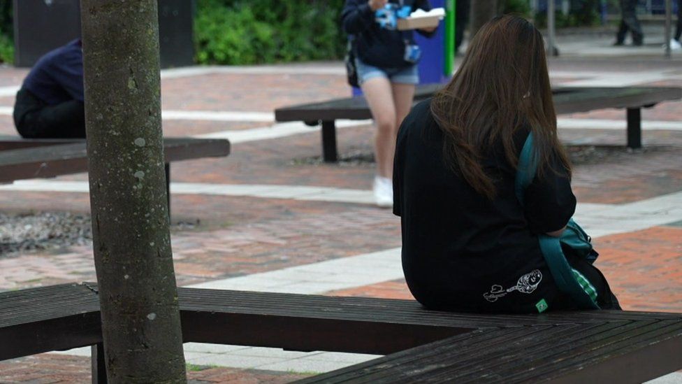 Image of an anonymous student sitting on a bench in a square on a university campus.