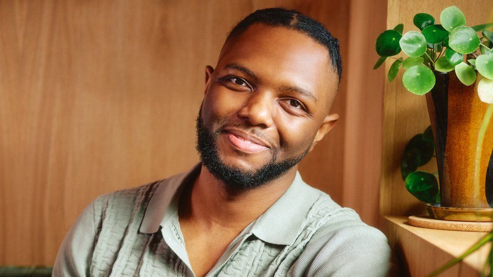 A young black man with short, black hair and beard sits in a wood-panelled room. He's sitting, leaning slightly against a chair or sofa back. He smiles warmly, illuminated by the light from the neighbouring window which casts a smooth swathe of light across the room.
