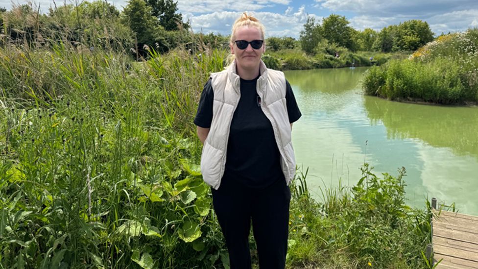 A smiling woman in a black top and t-shirt, with a cream body warmer over the top. She is wearing sunglasses and standing by the water at a fishing lake
