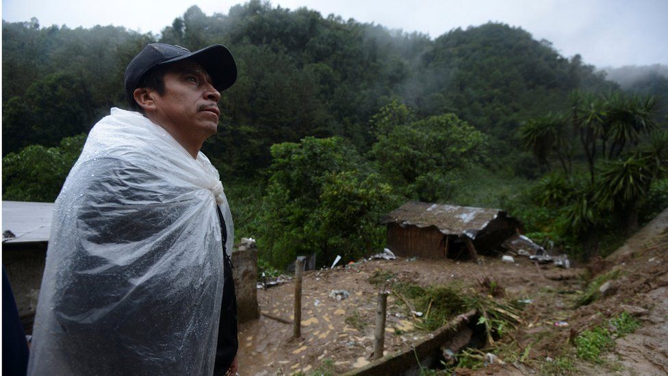 man wrapped in plastic sheeting to protect himself from rain, near destroyed house on muddy hillside, 7 August 2016