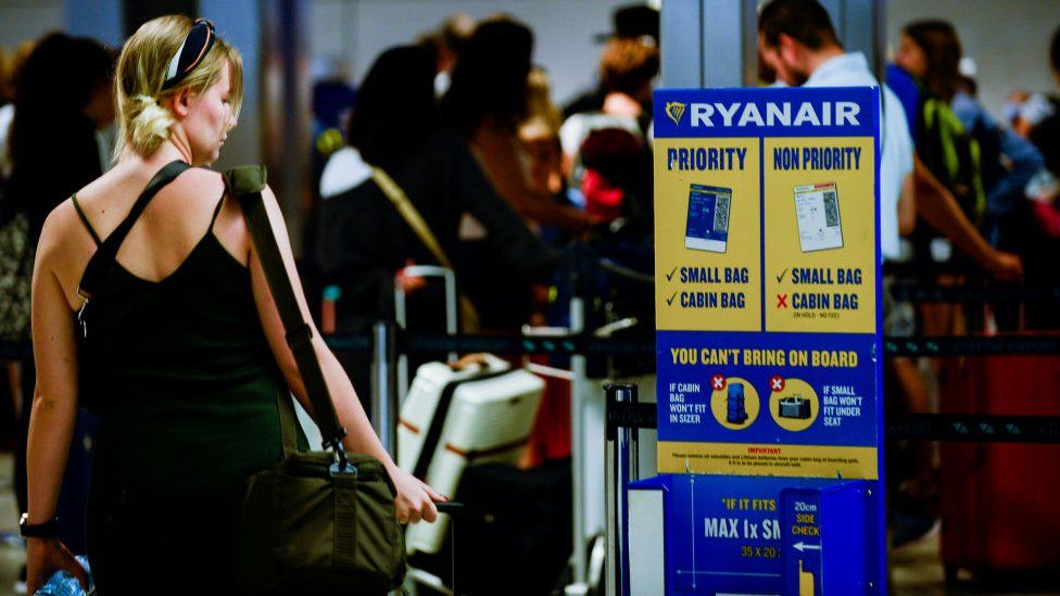 A blonde woman passes by a Ryanair luggage information stand at an airport. The luggage stand is blue and yellow and details what can be brought on board and the permitted baggage dimensions