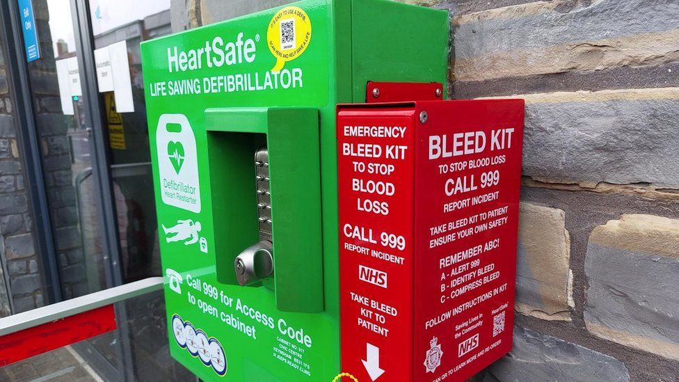 A green defibrillator on a wall. A red bleed kit box has been installed next to it. It reads "Emergency bleed kit to stop blood loss" with the 999 emergency number and NHS logo underneath in white letters.