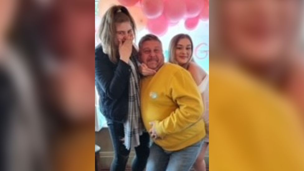 Paul Morton in a yellow hoodie being hugged by two young women. One has her hand over her mouth.