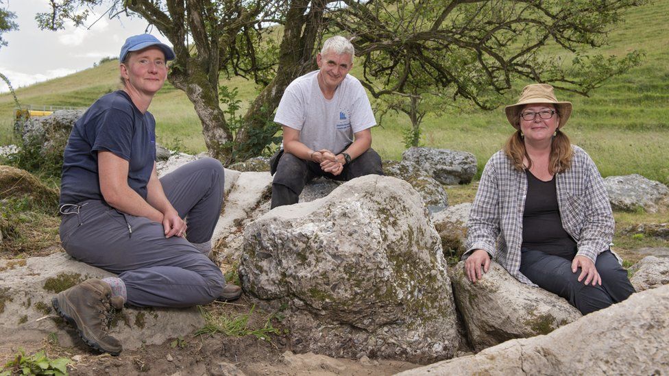 two men and a woman sat by a boulder in the countryside