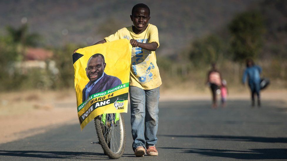 A boy pushes his bicycle as he holds a candidate poster of the South African ruling party African National Congress on the eve of South African Municipal elections on August 02, 2016 in Vuwani