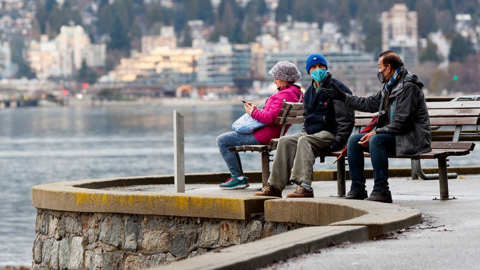 Two man wearing protective face mask converse on a park bench along the seawall at Stanley Park