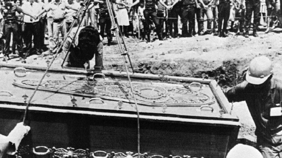 A black and white photo showing a coffin being lowered into a grave in 1987