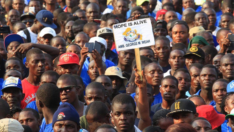 a crowd of faces, and one sign that reads "Hope is Alive George Weah"