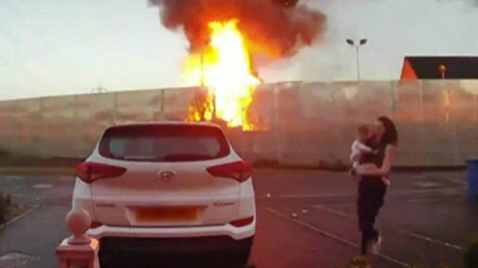 A fire takes hold in a fence, as a woman carrying a baby runs to a white car parked in a car park, opposite the fire