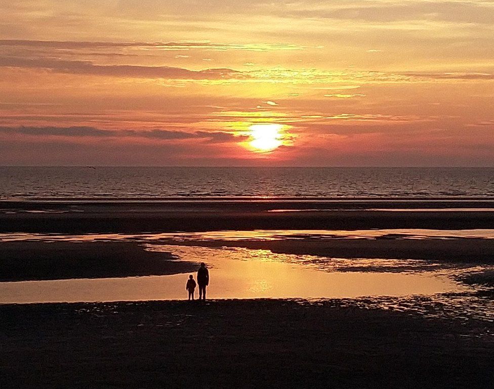 An adult and a child stand on a beach looking at the sea as the sun sets